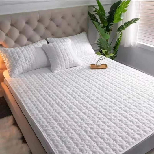 White quilted mattress protector fitted onto a bed with a light-colored headboard, with two matching pillows and a wooden tray on the bedside table.