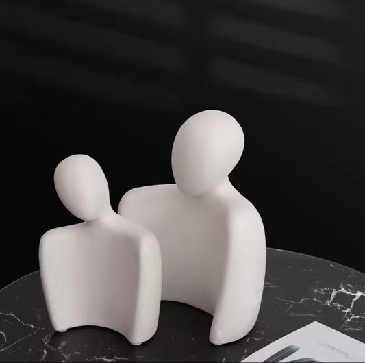 Two abstract, faceless white sculptures depicting a leaning couple, positioned on a black marble patterned table with a dark background.
