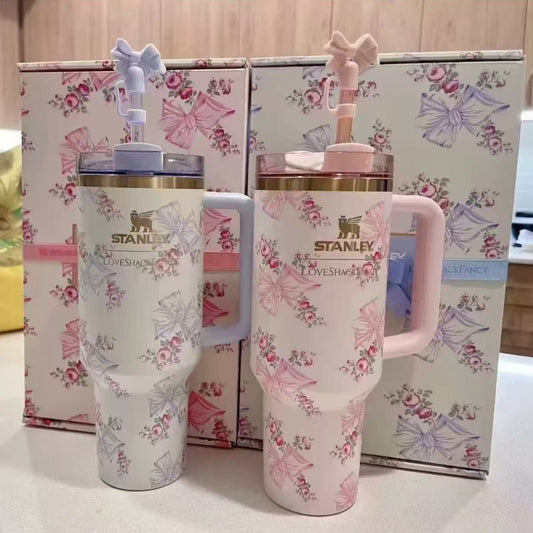 Two large Stanley brand insulated cups with handles, featuring a pink, blue, and white floral and bow pattern, one with a blue handle and one with a pink handle, standing in front of matching patterned gift boxes.