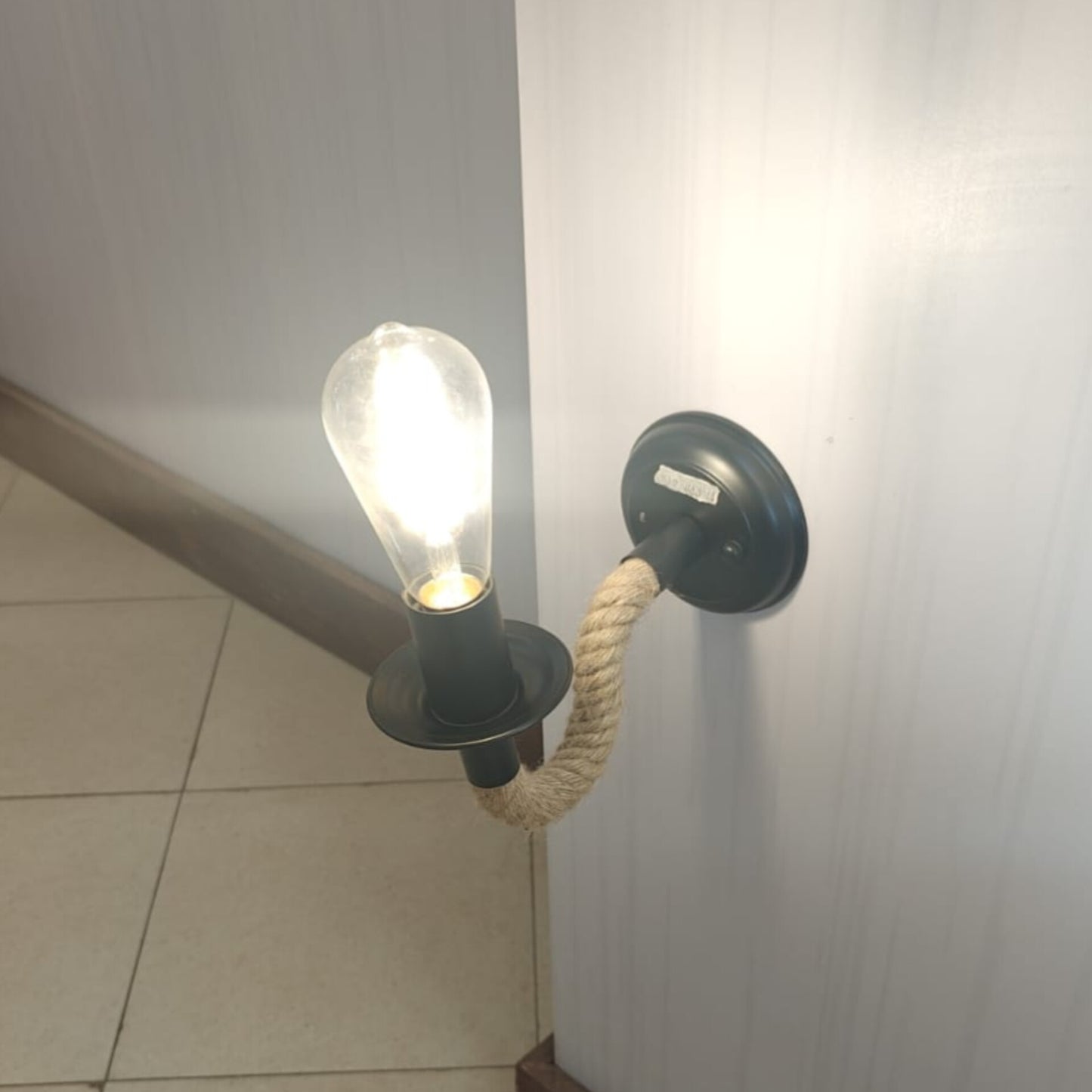A black wall mounted light fixture with a thick braided rope arm holding a lit clear glass bulb.