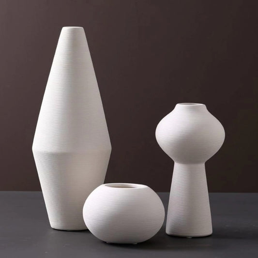 A set of three modern white ceramic vases with a subtle horizontal ribbed texture: one tall conical shape, one spherical shape, and one short pedestal shape, displayed on a dark surface.
