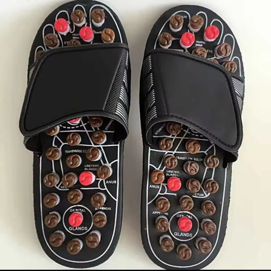 A pair of black reflexology massage slippers with numerous brown and red plastic massage nodes and a foot map diagram printed on the footbed, showing corresponding body areas.