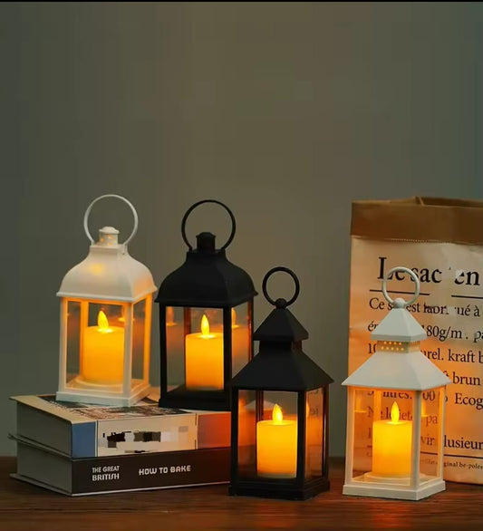 Four small decorative lanterns (two black, two white) with peaked tops and carrying handles, each containing a battery operated LED candle with a warm, flickering light.