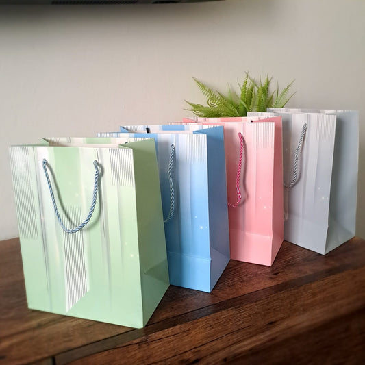 Four beautiful pastel gift bags in green, blue, pink, and gray with braided handles, sitting on a wooden table.