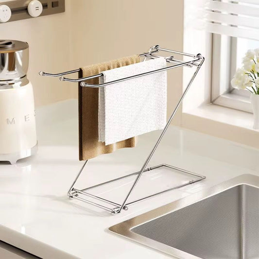 A polished silver stainless steel countertop towel rack with a Z shaped frame, holding two folded kitchen towels, one white and one brown, next to a kitchen sink