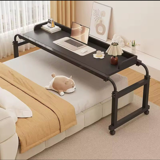 A black adjustable rolling desk placed over a white bed, holding a silver laptop, a coffee cup, and two framed art pieces, with the desk legs visible on both sides.
