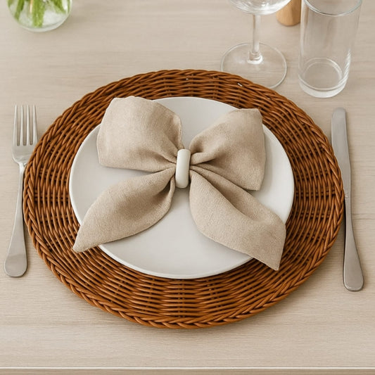 A single brown woven rattan style round place mat set with a white plate, a beige linen napkin tied in a bow, and silverware on a light wooden table.