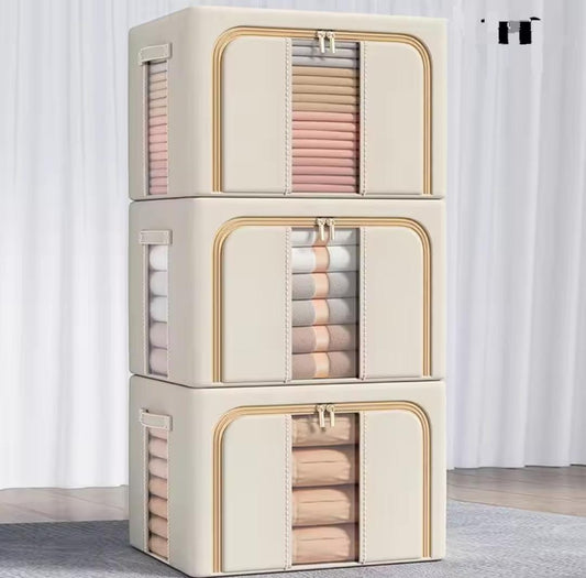 Three stacked large, beige fabric storage organizer boxes with clear front windows and gold accents, showing neatly folded blankets and duvets inside.