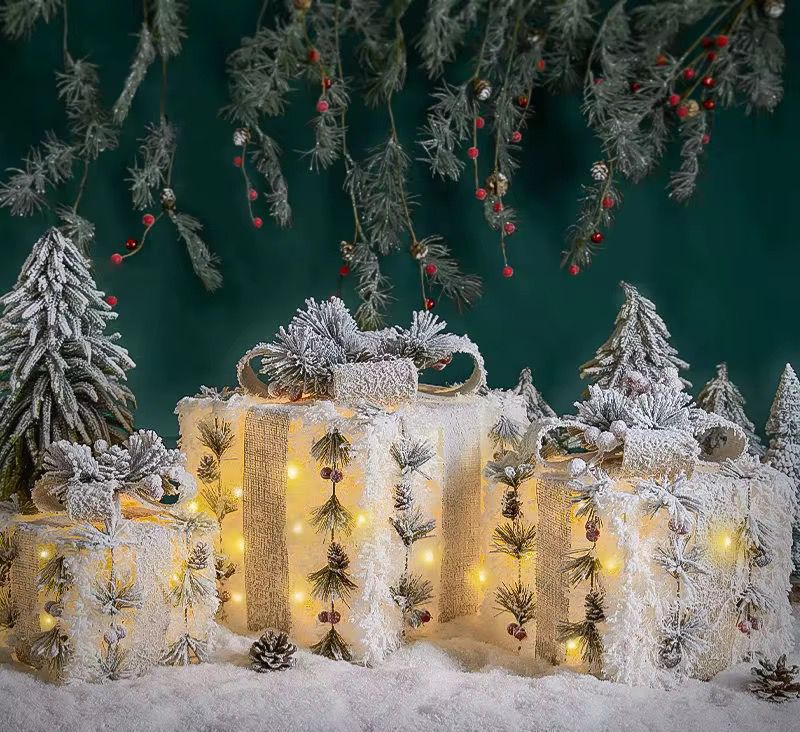 Three snow-flocked lighted Christmas gift boxes decorated with pine, berries, and a burlap texture