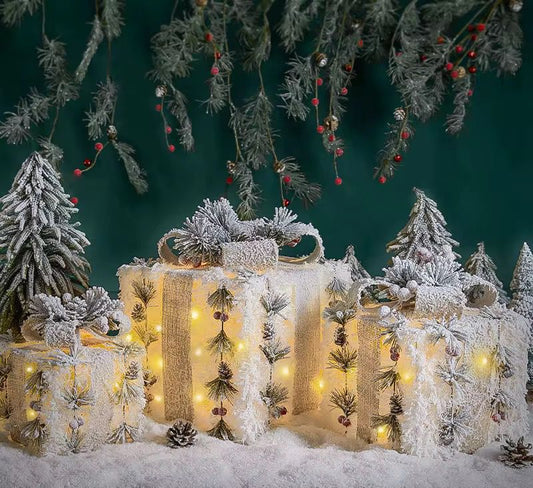 Three snow-flocked lighted Christmas gift boxes decorated with pine, berries, and a burlap texture