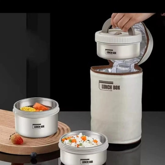 Three stainless steel round lunch boxes with lids, with one being placed into a tall, beige insulated lunch box bag on a dark background.