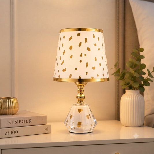 A table lamp with a white drum shade decorated with gold speckles, a gold-finished metal base, and a faceted clear crystal-like pedestal foot.