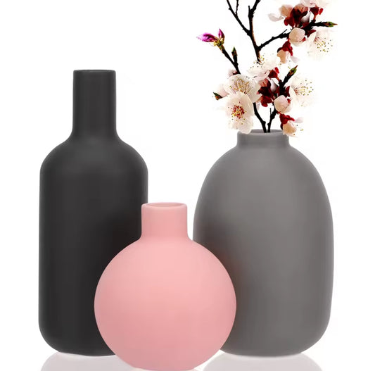 A set of three modern ceramic vases in muted colors: a tall dark gray bottle shape, a small pink sphere, and a medium gray oval shape holding white and pink flowering branches, set against a white background.