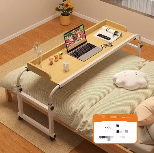 A light wood and white adjustable rolling over bed desk with a raised tray edge, holding a laptop, coffee cup, and remote control, positioned over a bed with a white comforter.