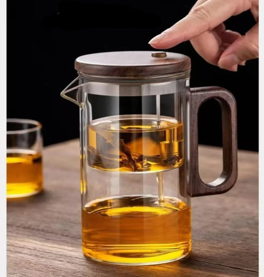 Clear glass tea infuser pot with a wood-look lid and handle, showing dark tea leaves steeping in the upper chamber and clear amber tea in the main pot below.