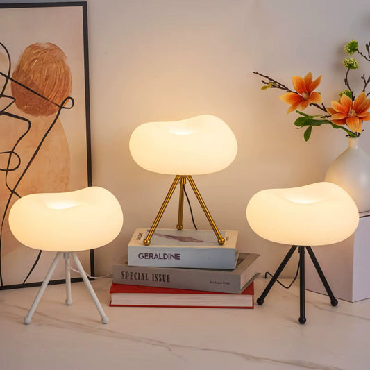 Three tripod cloud table lamps with different colors (white, gold, and black) positioned on a table with books and a flower vase in the background.