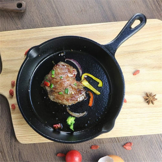 A black, round cast iron skillet holding a cooked steak with sliced peppers and onions, resting on a wooden cutting board with spices visible around the edge.