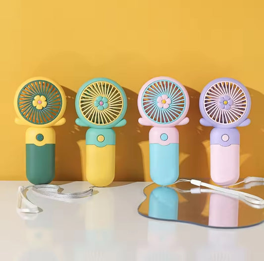 Four colorful mini handheld portable fans with flower blade guards lined up against a yellow wall, showing the different color combinations (green yellow, blue yellow, pink blue, and purple pink).