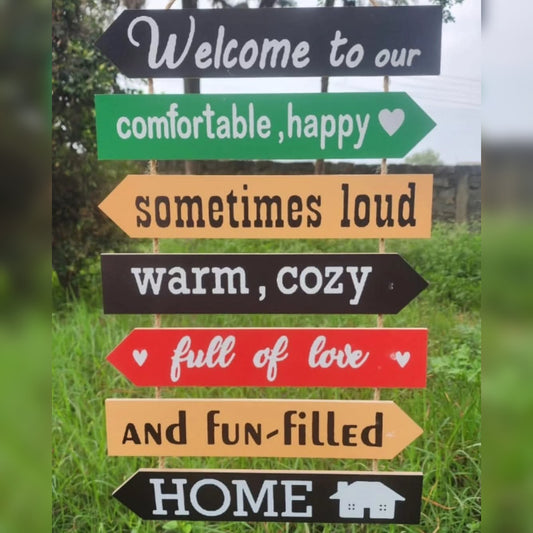 A vertical stack of six colorful, arrow shaped wooden signs hung by twine against a green background, listing rules and welcome phrases for a home.