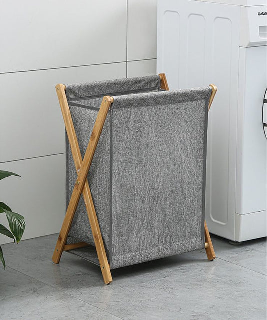 A tall, gray fabric laundry hamper with a large capacity, supported by a natural wooden bamboo folding X frame, positioned next to a white washing machine.