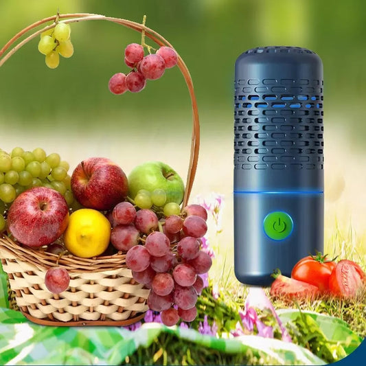 A dark blue portable cylindrical fruit and vegetable food cleaner next to a basket of fresh grapes, apples, and lemons outdoors.
