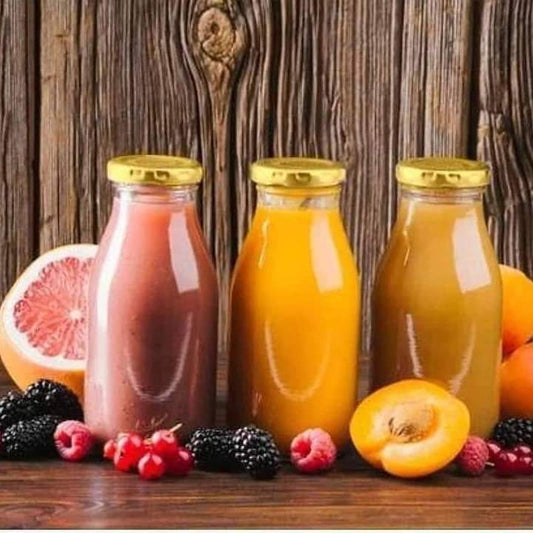 Three glass bottles filled with pink and orange juice standing on a wooden surface next to various fruits like grapefruit, berries, and a sliced apricot, all topped with shiny golden lids.